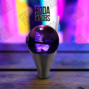 May include: A custom shift knob with a clear, spherical top featuring a purple, glowing character. The knob is mounted on a silver-colored base. The text "SODA KNOBS" is displayed above the knob.