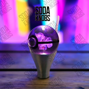 Könnte beinhalten: Ein klarer, kugelförmiger Soda Knobs Schalthebelknauf mit einer lila, beleuchteten Kreatur im Inneren. Der Knauf sitzt auf einem silbernen Sockel, mit dem Soda Knobs-Logo in Schwarz darüber. Der Hintergrund ist ein verschwommenes Lila, Gelb und Blau.