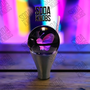 Peut inclure: Un bouton de soda décoratif avec un dessus sphérique transparent orné d'un motif de papillon rose. Le bouton a une base argentée et le texte "SODA KNOBS" est affiché au-dessus en noir.
