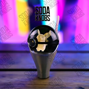 Puede incluir: Una esfera de vidrio negro y transparente con un diseño de Pikachu, montada sobre una base plateada. El texto "SODA KNOBS" está encima. El diseño de Pikachu es amarillo. La esfera está sobre una superficie de madera.
