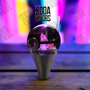 Puede incluir: Un pomo de cambio personalizado con una parte superior esférica transparente con un diseño geométrico rosa. El pomo tiene una base plateada y el texto "SODA KNOBS" se muestra en negro.