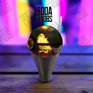 Peut inclure: Un bouton de soda personnalisé avec un dessus sphérique transparent présentant un motif de grenouille coloré et éclairé. Le bouton a une base argentée et le texte "SODA KNOBS" est affiché en noir.