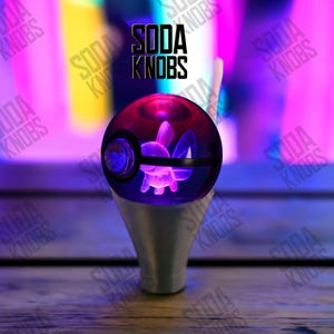 Op de afbeelding: Een aangepaste sodaknop met een helder, bolvormig ontwerp en een paars interieur, met een zwarte band. De knop heeft een zilveren basis en de tekst "SODA KNOBS" wordt hierboven weergegeven.