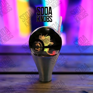 Peut inclure: Un bouton de soda noir et argenté avec un personnage de dessin animé lumineux à l'intérieur d'une sphère transparente. Le texte "SODA KNOBS" est affiché au-dessus du bouton. L'arrière-plan est un mélange flou de violet, de jaune et de bleu.