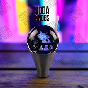 Könnte beinhalten: Ein schwarzer und silberner Schalthebelknauf mit einer transparenten, kugelförmigen Oberseite. Im Inneren der Kugel befindet sich ein blau-weißes Design. Der Text "SODA KNOBS" wird in schwarzen Buchstaben über dem Knopf angezeigt.