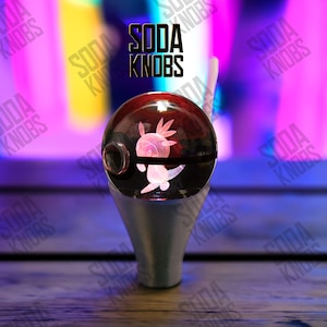 Könnte beinhalten: Ein klarer, runder Schaltknauf mit roter Oberseite und silberner Basis. Der Knauf zeigt ein leuchtendes rosa Axolotl-Design. Der Text "SODA KNOBS" ist in Schwarz über dem Knauf angezeigt.