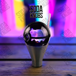 May include: A black and silver soda knob with a clear, spherical top. The top has a purple design inside. The base is silver and cone-shaped. The text "SODA KNOBS" is displayed in black.