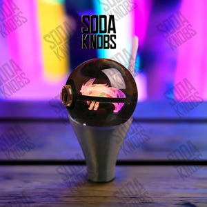Könnte beinhalten: Ein individueller Schalthebelknauf mit einer transparenten, kugelförmigen Oberseite mit einem rosa-weißen Tierdesign. Der Knauf hat einen silbernen Sockel und der Text "SODA KNOBS" ist im Hintergrund sichtbar.
