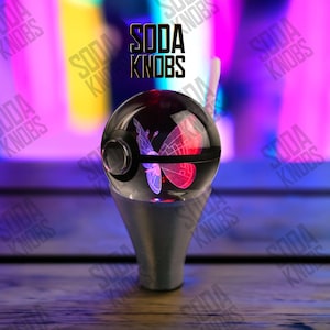 Könnte beinhalten: Ein klarer, kugelförmiger Soda-Knauf mit einem Schmetterlingsdesign, das rote und violette Farbtöne aufweist. Der Knauf sitzt auf einem silberfarbenen Sockel. Der Text "SODA KNOBS" wird über dem Knauf angezeigt.