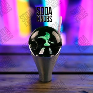 May include: A decorative soda knob featuring a green figure within a clear glass orb, mounted on a silver-toned base. The text "SODA KNOBS" is visible above the orb. The background is a mix of purple and yellow.