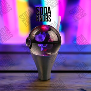 Pode incluir: Um botão de refrigerante esférico transparente com um design de personagem roxo no interior. O botão fica em cima de uma base prateada. O texto "SODA KNOBS" é exibido em preto acima do botão.