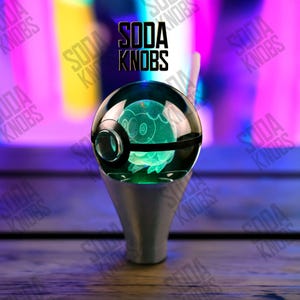 Op de afbeelding: Een aangepaste versnellingspookknop met een heldere, bolvormige bovenkant met een gloeiend groen ontwerp. De knop heeft een zwarte band en een zilveren basis. De tekst "SODA KNOBS" staat erboven.