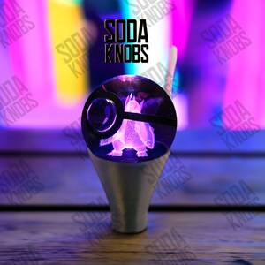 Puede incluir: Un pomo de soda personalizado con un pingüino morado brillante dentro de un globo transparente con un anillo negro. El pomo está sobre una base plateada. El texto "SODA KNOBS" se muestra arriba.