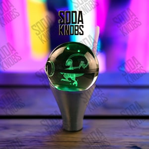 Peut inclure: Un bouton de soda vert et brillant sur le thème des dinosaures avec le texte "SODA KNOBS" en noir. Le bouton est une sphère transparente avec une silhouette de dinosaure à l'intérieur, montée sur une base argentée. L'arrière-plan est un mélange flou de violet et de bleu.