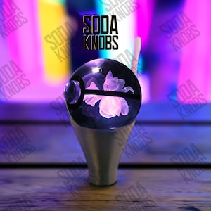 Könnte beinhalten: Ein individueller Schaltknauf mit einer transparenten, kugelförmigen Oberseite, die ein leuchtendes rosa Blumendesign umschließt. Der Knauf hat einen silbernen Sockel und der Text "SODA KNOBS" wird darüber angezeigt. Der Hintergrund ist eine verschwommene Mischung aus Lila und Gelb.