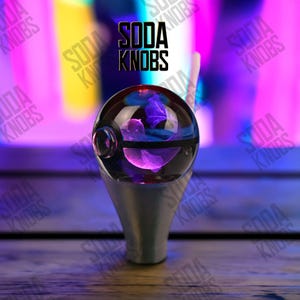 Peut inclure: Un bouton de soda décoratif avec un dessus sphérique transparent contenant des accents violets et bleus. Le bouton est monté sur une base argentée. Le texte "SODA KNOBS" est affiché en noir.