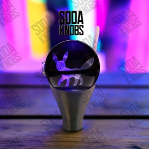 Peut inclure: Pommeau de levier de vitesse sphérique transparent avec un design Umbreon violet brillant. Le pommeau est monté sur une base argentée. Le texte "SODA KNOBS" est affiché au-dessus du pommeau.