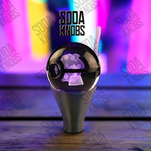 Peut inclure: Un objet sphérique noir et transparent avec un motif de champignon, monté sur une base argentée. Le texte "SODA KNOBS" est affiché au-dessus de la sphère. Le fond est un dégradé de violet et de jaune.