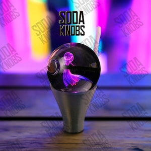 Könnte beinhalten: Ein klarer, kugelförmiger Soda-Knopf mit einem lila und rosa beleuchteten Design im Inneren, montiert auf einem silberfarbenen Sockel. Der Text "SODA KNOBS" wird in Schwarz über dem Knopf angezeigt.