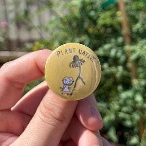 May include: A round, yellow button with the words "PLANT NATIVE" in black, curved text. The button features a black illustration of a flower and a small figure. The button is held in a hand against a blurred green background.