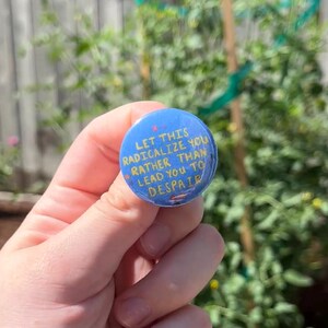 May include: A blue button with the text "LET THIS RADICALIZE YOU RATHER THAN LEAD YOU TO DESPAIR" in yellow and white. The button is held in a hand against a blurred green background.