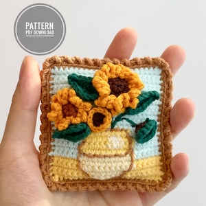 May include: Crocheted sunflower artwork in a brown frame. The sunflowers are yellow with brown centres and green leaves. The artwork is on a light blue background with a yellow and brown base.
