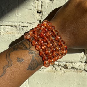 Puede incluir: Una pila de pulseras de cuentas transparentes teñidas de naranja. Las pulseras están hechas de grandes cuentas redondas ensartadas con espaciadores dorados. Las pulseras se usan en una muñeca con un tatuaje visible en el brazo.