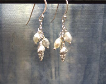 May include: A pair of dangle earrings featuring small, white pearls and silver seashell charms. The earrings hang from silver hooks and are displayed against a blue background. The earrings are a delicate and elegant accessory.