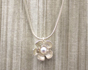 May include: A silver-toned necklace featuring a flower pendant with a central pearl. The flower pendant has detailed petals. The chain is a thin, snake-style design. The jewelry is displayed against a neutral background.