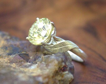 May include: A gold ring with a large, faceted yellow gemstone. The ring features a leaf design on the band, adding a natural and elegant touch. The gemstone is held in place by delicate prongs.