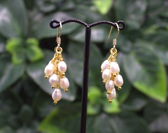 May include: A pair of elegant gold-toned earrings featuring clusters of small, creamy white pearls. The pearls are suspended from delicate gold wires, creating a cascading effect. The earrings are displayed against a blurred green background.