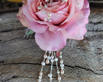 May include: A pink rose hair comb with a silver wire and pearl accent. The comb features a large, soft pink rose with a delicate pearl and wire design. Pearl strands cascade from the rose, creating an elegant, romantic look.