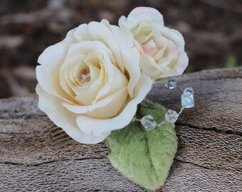 May include: A close-up of a floral hair accessory featuring two ivory-colored roses with a touch of pink, a green leaf, and a silver wire adorned with clear, iridescent beads. The roses have detailed petals.