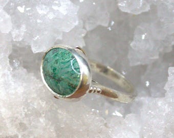 May include: A silver ring featuring a round, vibrant green stone with dark green marbling. The ring has a simple band with small decorative details near the stone. The gemstone is set in a silver bezel.