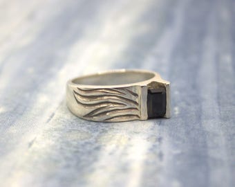 May include: A silver ring with a rectangular black gemstone. The ring band features a textured wave design on the side. The gemstone is set in a square bezel setting, creating a modern and bold look. The ring is displayed on a blue and white patterned surface.