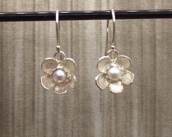 May include: A pair of silver-colored flower earrings with a pearl center. The earrings are hanging from a black bar against a neutral background. The flower petals have a textured finish.
