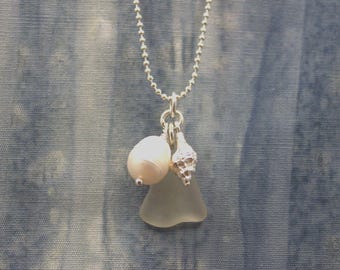 May include: A silver necklace with a pendant featuring a white pearl, a small seashell, and a piece of sea glass. The necklace has a delicate chain and is set against a blue background.
