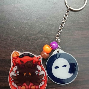 May include: A Halloween-themed keychain with two round charms. One charm features a red and orange illustration of a creature with a pumpkin and ghosts. The other charm is blue with a white ghost silhouette. The charms are connected by a silver chain with colorful beads.