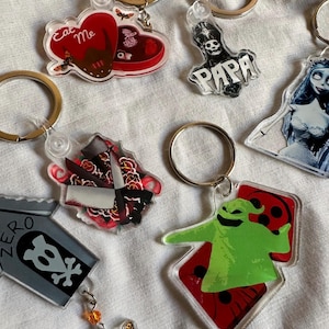 May include: A collection of acrylic keychains with various designs. Keychains include a red heart with "Eat Me" text, a skull with "PAPA" text, a knife with floral accents, a tombstone with a skull, and a green character on dice.