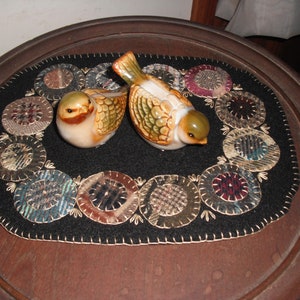 May include: A black felt table runner with a circle design featuring a variety of fabrics in shades of brown, blue, and white. Two ceramic birds with brown, green, and white accents are placed on the runner.