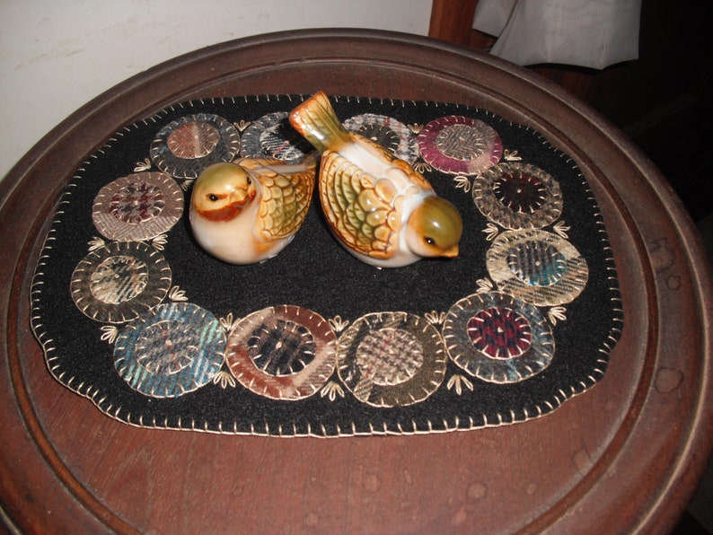 May include: A black felt table runner with a circle design featuring a variety of fabrics in shades of brown, blue, and white. Two ceramic birds with brown, green, and white accents are placed on the runner.