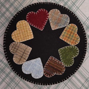 May include: A black felt coaster with a circle of ten heart-shaped patches in various colors and patterns. Each heart is stitched with white thread. The coaster is a decorative item.