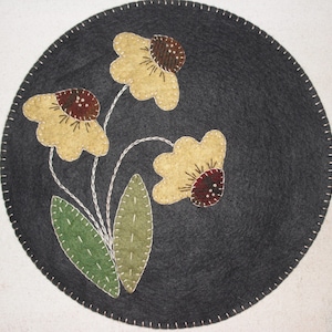 May include: A round, gray felt rug with a stitched border features three yellow flowers with brown centers and green leaves. The flowers are stitched in a simple, folk art style.