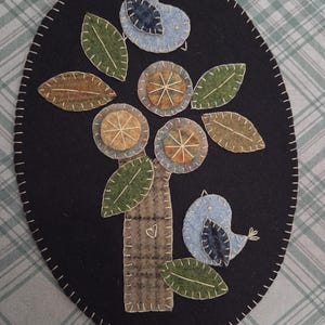 May include: Oval-shaped, black felt applique featuring a tree with circular fruit and green leaves. Two blue birds perch on the tree. The tree trunk has a heart. The edge is stitched with white thread. A charming, handmade piece.