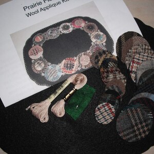 May include: A wool applique kit for a prairie flower design. The kit includes a black felt base, a variety of patterned wool fabric circles, two spools of embroidery floss, and a green felt piece.