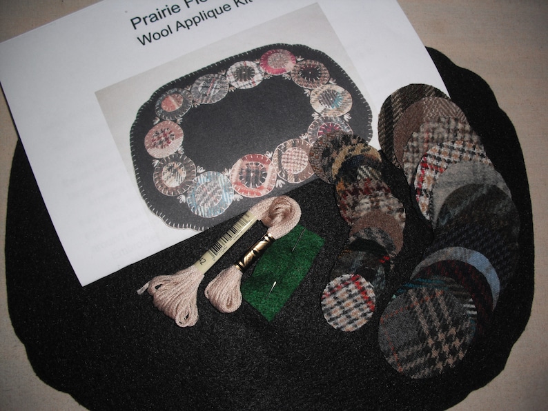May include: A wool applique kit for a prairie flower design. The kit includes a black felt base, a variety of patterned wool fabric circles, two spools of embroidery floss, and a green felt piece.