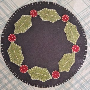 May include: A round, black felt coaster with a white stitched border. The coaster is decorated with green felt holly leaves and red felt berries, creating a festive holiday design. The coaster is placed on a blue and white plaid surface.