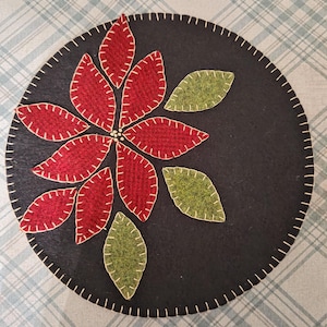 May include: A round, black felt trivet with a poinsettia design. The poinsettia has red petals with white stitching, and green leaves. The edge is finished with white stitching. The trivet is on a blue and white plaid surface.