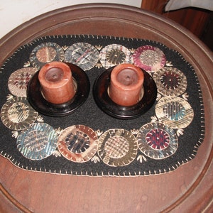 May include: A black felt table runner with a circular design of various fabrics in shades of brown, beige, blue, and red. Two brown candle holders with brown candles are placed on the runner.