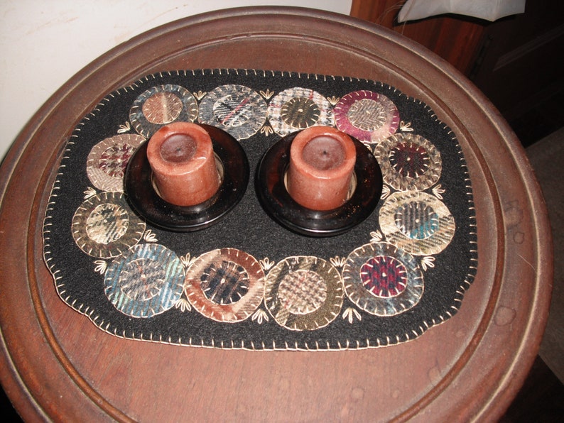 May include: A black felt table runner with a circular design of various fabrics in shades of brown, beige, blue, and red. Two brown candle holders with brown candles are placed on the runner.